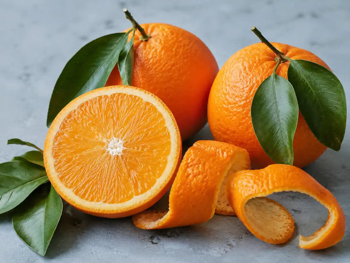 Fresh sweet oranges, a sliced orange and curled peel with glossy green leaves on a pale blue-grey background