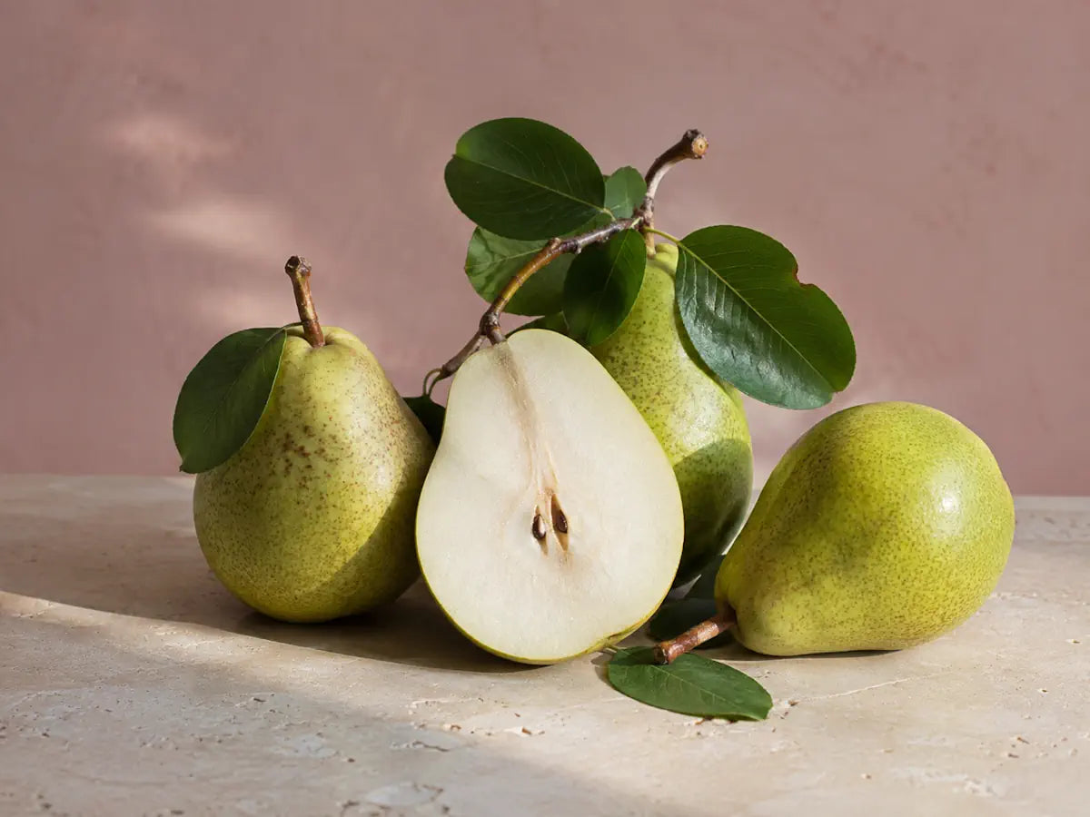 Fresh green pears, one sliced in half, with glossy leaves on a light dusty pink background