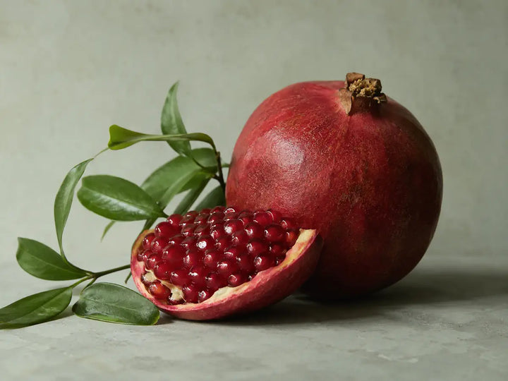 Open red pomegranate with ruby seeds and green leaves on a pale sage-grey background