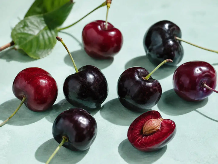 Deep red and dark cherries, one sliced to show the stone, with green leaves on a pale mint-blue background