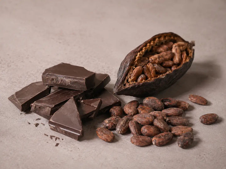 Dark chocolate pieces, roasted cocoa beans and a broken cacao pod on a soft rose-grey background