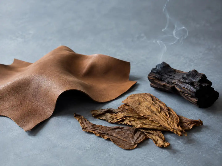 Natural leather, dried tobacco leaves and lightly charred wood on a pale smoky blue-grey background