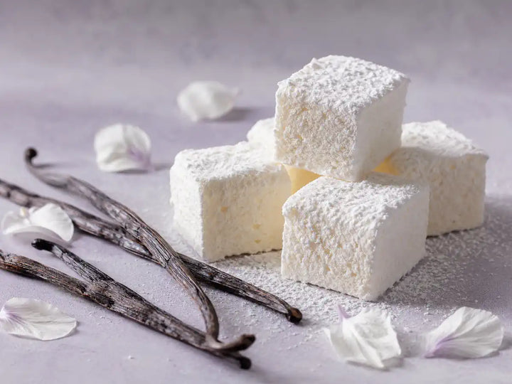 White marshmallows with vanilla pods and pale petals on a lavender-grey background