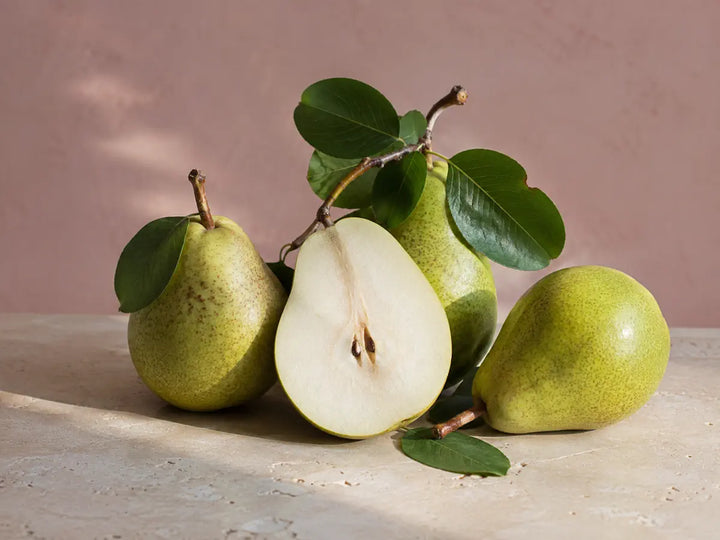 Fresh green pears, one sliced in half, with glossy leaves on a light dusty pink background