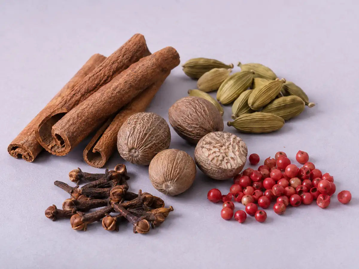 Cinnamon bark, cardamom pods, nutmeg, cloves and pink peppercorns on a soft lavender-grey background